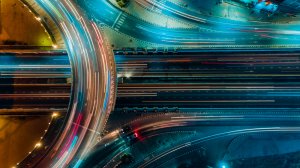Aerial view of a futuristic highway interchange illuminated with blue and yellow lights. The infrastructure is busy with streaks of light from fast-moving vehicles, creating a dynamic and vibrant scene at night. The layout showcases multiple lanes and curved paths.