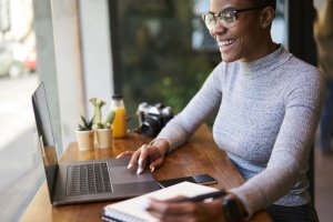 A person wearing glasses and a gray turtleneck sits at a wooden table, remotely working on a laptop. They are smiling and writing in a notebook. Nearby are small potted plants, a bottle of orange juice, and a camera. A window provides natural light.