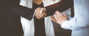 Two individuals in suits are shaking hands firmly, embodying leadership. Another person, dressed in white, stands nearby with their hands clasped together. The background is blurry, focusing the attention on the handshake, symbolizing an agreement or meeting.