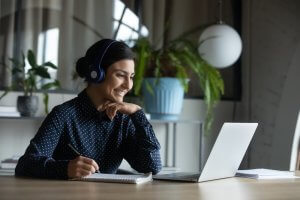 A person wearing headphones is sitting at a table, smiling and taking notes while looking at a laptop. Engaged in remote working, the scene includes a large potted plant and light curtains in the background.