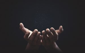 A pair of cupped hands are held up against a dark background, illuminated softly from above. Small, glowing particles float above the hands, creating a sense of wonder and the spark of life.