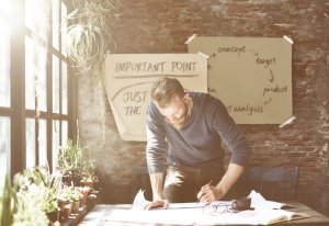 A bearded man is leaning over a table in a sunlit room, immersed in his work with productivity. Behind him, a brick wall displays handwritten notes on papers taped to it. The table is adorned with several small potted plants and a coffee cup.
