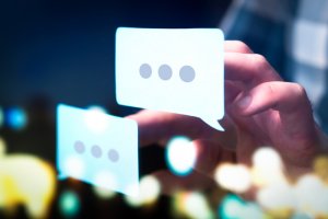 A person's hand is interacting with two digital speech bubbles, each containing three dots, symbolizing typing or messaging. The background features a blurred, colorful array of lights and shapes, suggesting a digital work or online communication context.