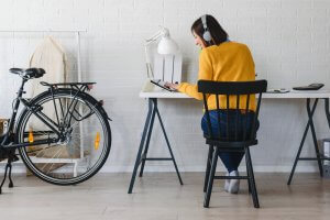 A person wearing headphones and a yellow sweater sits at a desk focusing on a tablet, embracing the flexible work environment. The workspace features a table lamp, a bicycle leaning against a white brick wall, and a wooden chair. In the background, there is a cloth cover on a clothing rack.