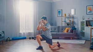 A man is performing a lunge exercise in a spacious, well-lit living room, aiming to get in shape. The room has a gray sofa, wooden floor, and light-colored walls adorned with framed pictures. Fitness equipment like a yoga mat and dumbbells are visible in the background.