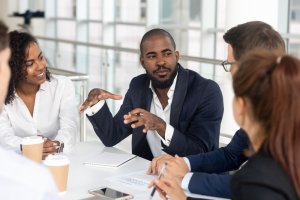 A diverse group of four people are sitting at a table in a modern office, engaged in an animated discussion. One man, wearing a dark suit, is speaking and gesturing with his hands, making the meeting more effective while the others listen attentively. Coffee cups and notepads are on the table.