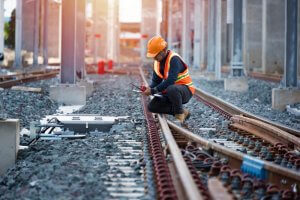 A construction worker wearing a hard hat and high-visibility vest works on railroad tracks, contributing to essential infrastructure. Kneeling, they examine or adjust equipment with tools. The surrounding area shows a construction site with gravel and structural elements.