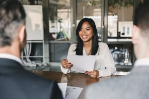 A woman sits at a table, smiling and holding a resume. She is facing two men in suits, with their backs to the camera. The scene appears to be an interview or a professional meeting in a modern office setting.