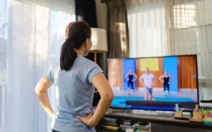 A woman stands in her living room, facing a television screen that displays an exercise video featuring three instructors. She is wearing workout clothes, ready to get in shape, with her hands on her hips. The room is bright with sunlight streaming through sheer curtains.
