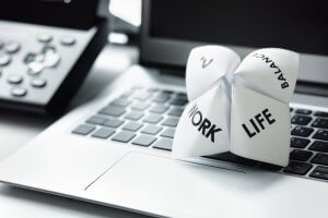A paper fortune teller labeled with "Work" on one flap and "Life Balance" on another flap sits on a laptop keyboard, underscoring the importance of flexible work and maintaining balance. In the background, a phone subtly reminds us of continuous connectivity.
