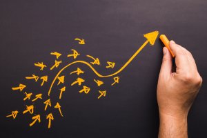 A person's hand is holding an orange piece of chalk and drawing an upward-curving arrow on a chalkboard. Around the larger arrow, numerous smaller arrows point in various directions but converge towards the main arrow's path, making the overall message more effective.