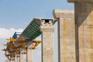 A concrete bridge is under construction, with large pillars supporting steel frameworks. A segment of the bridge deck, partially covered with a green net, extends across the supports. Clear blue sky is visible in the background, highlighting this vital piece of infrastructure.
