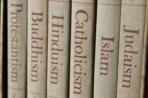 Close-up of six book spines, each labeled with a different religion: Protestantism, Buddhism, Hinduism, Catholicism, Islam, and Judaism. The titles are printed in red on a beige, textured fabric cover, offering an insight into the diverse philosophies that shape human life.