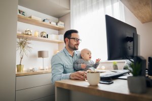 A man with glasses sits at a desk working on his computer while holding a baby on his lap, embodying the essence of remote working. The baby looks towards the screen, appearing engaged. The desk is situated in a well-lit room with shelves holding decorative items and books in the background.