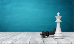 A chess king piece stands upright in triumph, while a queen piece lies on its side, defeated, on a light wooden surface. The background is a smooth teal gradient wall, highlighting the winners and losers of this strategic battle.