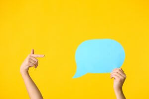 Two hands are shown against a bright yellow background. One hand on the left is pointing, while the hand on the right is holding a large light blue speech bubble cutout filled with quotes. The image suggests communication or expression.