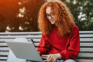 A young person with curly red hair and glasses is sitting on a bench, using a laptop. They are wearing a red sweatshirt and are focused on their screen, possibly looking for a job. A backpack is beside them. The background shows green trees, suggesting a park or outdoor area.