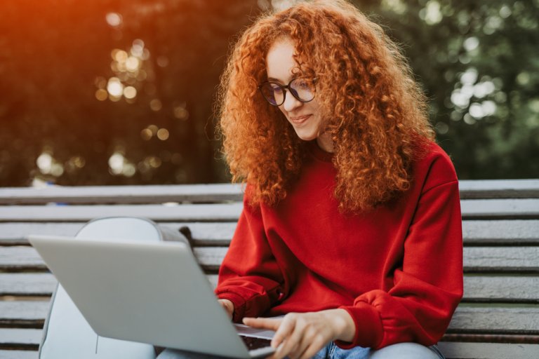 A young person with curly red hair and glasses is sitting on a bench, using a laptop. They are wearing a red sweatshirt and are focused on their screen, possibly looking for a job. A backpack is beside them. The background shows green trees, suggesting a park or outdoor area.