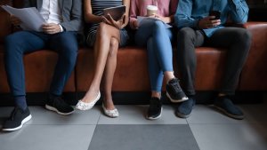 Four people sitting on a brown leather couch, dressed in business casual attire, each holding a different item: papers, a tablet, a coffee cup, and a phone. Only their lower halves are visible, focusing on their legs and footwear—capturing the essence of job readiness.