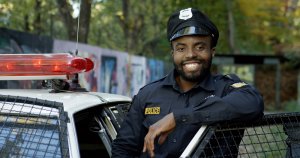 A smiling police officer in uniform stands next to a police car with the driver's side door open and a red siren light on top. The background features graffiti-covered walls and trees with autumn foliage, showcasing the vibrant community that the Police diligently serve and protect.