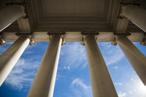 A low-angle view of tall white columns extends upwards against a vibrant blue sky with scattered white clouds, akin to the meticulous presentation of a federal resume. The composition emphasizes both the grandeur and architectural detail of the columns.