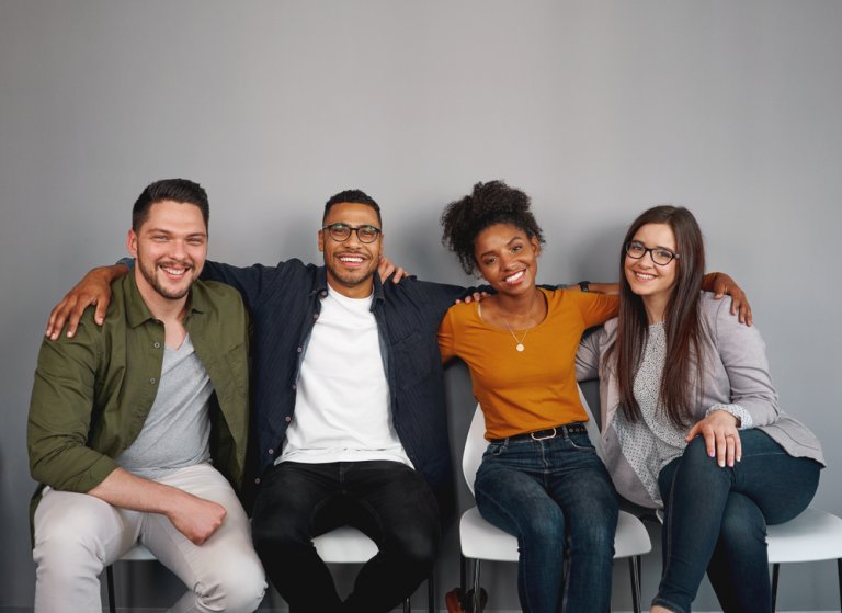 A group of four diverse people sit close together on chairs, smiling at the camera. They have their arms around each other, suggesting camaraderie and friendship. The background is a plain gray wall, highlighting their unity and commitment to DEI principles.