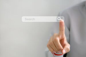 A person is pressing a transparent search button on a virtual interface, looking for job opportunities. The word "Search" is displayed in a search bar with a magnifying glass icon. The background is blurred and neutral gray, focusing on the person's hand and finger.