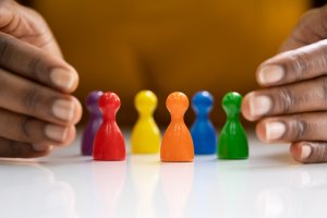 Close-up of hands encircling seven colorful wooden pawns arranged in a semicircle on a white surface, symbolizing DEI. The pawns are painted in red, yellow, purple, blue, orange, green, and another yellow. The background is blurred and features a warm, golden hue.