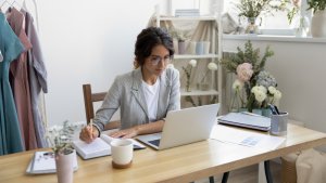 A person with glasses works on a laptop at a wooden desk, writing in a notebook. The workspace, an ideal spot for a creative job, features a coffee mug, color swatches, organized binders, and various potted plants and flowers, creating a bright and serene atmosphere. Decorative clothing hangs to the side.