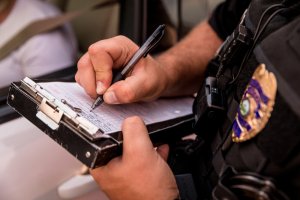 A close-up of a police officer's hands writing a ticket on a clipboard. The police officer is wearing a uniform, and a badge is visible on the right side of the image. The officer is standing next to a car with a person visible in the driver's seat.