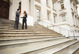 Two business professionals, a man and a woman, stand on the wide stone steps of a grand, classical-style building engaged in conversation. Both are dressed in formal attire, holding folders that contain their federal resumes. The building features tall columns and large windows.