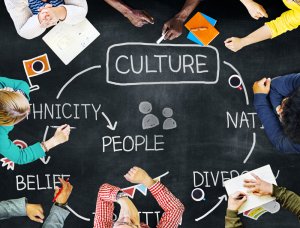 A group of diverse people sit around a chalkboard table, engaged in discussion. The table is filled with notes, books, and coffee cups. Words on the table include "CULTURE," "PEOPLE," "ETHNICITY," "BELIEF," and "DIVERSITY," connected by arrows, illustrating their vibrant work culture.
