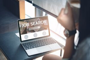 A person sits at a desk looking at a laptop screen displaying the words "JOB SEARCH" with a search bar and icons. Holding a cup, they seem to be taking a break or focusing on their job hunt in this casual, well-lit environment—their pursuit for the perfect job continues.