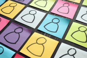 A collection of colorful sticky notes, each featuring a simple stick figure drawing of a person, arranged in a grid on a dark surface. The sticky notes are in various colors including yellow, pink, blue, green, and white—showcasing the essence of DEI through diverse and inclusive visual representations.