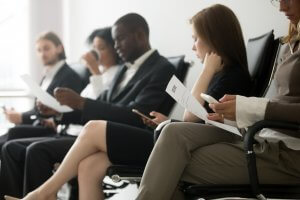 A group of five individuals sits in a waiting room, each holding documents and looking thoughtful. Some are reading papers, likely for a job interview, while others are using their smartphones. All are seated in a row of black chairs, dressed in business attire, suggesting a professional setting.
