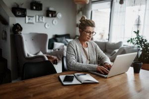 A person wearing glasses and a gray cardigan is sitting at a wooden table in a cozy living room, typing on a laptop, immersed in remote work. The table also has a notebook, smartphone, and a black mug. The background features a sofa, framed photos, and warm lighting.