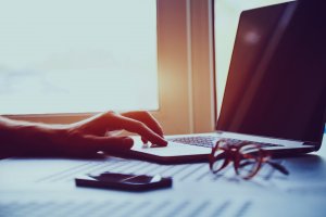 A person's hand is typing on a laptop keyboard in a dimly lit room, perhaps crafting a job description. A pair of glasses and a smartphone sit nearby on the table. Sunlight streams in through a window in the background, casting soft shadows across the scene.