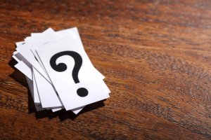 A small stack of white cards with a large black question mark printed on the top card rests on a wooden surface. The wood, rich in dark brown color with visible grain patterns, leads one to wonder about the mysteries the cards might hold.