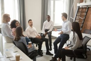 A group of seven diverse colleagues sit in a semi-circle in an office setting during a meeting. One person stands and talks animatedly while demonstrating leadership, while the others listen attentively, smiling and engaging. There are bright windows and modern office furniture in the background.