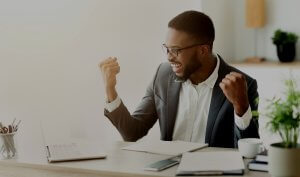 A man wearing glasses and a suit is sitting at a desk, raising both fists in a celebratory gesture. Smiling and looking at an open laptop showcasing video resumes, he is clearly pleased. Various office supplies and papers scatter the desk in the bright, modern office space.
