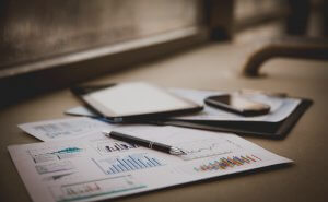 A desk with financial charts and graphs on papers, a pen, a tablet, and a smartphone placed on top, set near a window. The colorful bar and line graphs indicate accounting finance analysis in progress.