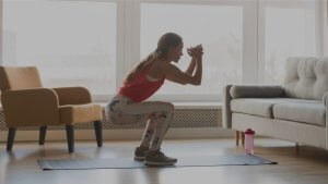 A person with long hair is performing a squat exercise on a yoga mat indoors, following their routine. They are in front of a large window, and in the background, there's a beige couch with a pink water bottle beside it and a mustard-yellow chair.