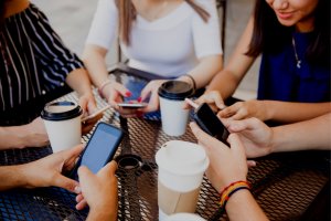 A group of Gen Z individuals sit at an outdoor table, each holding a smartphone and engaging with their devices. Several disposable coffee cups are on the table. They are casually dressed, and only their hands and parts of their torsos are visible.