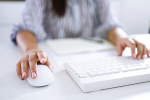 Close-up of a person using a computer mouse with one hand, while the other rests on a keyboard. They are seated at a white desk, and a notebook is visible in the background. The person, possibly drafting a job description, is wearing a long-sleeved shirt with stripes.