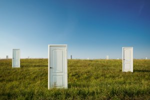 Three white doors stand independently in a green grassy field under a clear blue sky. The evenly spaced doors against the backdrop of an expansive horizon evoke a surreal and dreamlike atmosphere, inviting contemplation of choice.