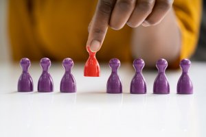 A hand is demonstrating leadership by picking up a red game piece from a group of six purple game pieces that are lined up on a white surface.