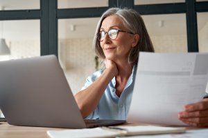 A woman with gray hair and glasses is sitting at a desk, looking at a laptop screen. She is holding a document in her hand and appears to be working or studying, possibly researching video resumes. There is a notebook and a pen on the desk in front of her.