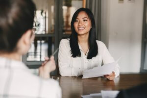 A woman with long black hair, wearing a white blouse, smiles and holds a document during an interview in a government office. She is seated at a table, facing another person who is holding a pen. The background shows a modern office setting with glass walls and a blurred figure.