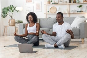 A man and woman sit cross-legged on yoga mats in a living room, meditating in front of a laptop as part of their daily routine. They both wear athletic clothes; the woman has on a white sports bra and grey leggings, and the man is dressed in a white t-shirt and grey sweatpants. A couch and shelves are in the background.