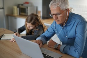 An older man with glasses wearing earphones focuses on his laptop, engaging in remote work, while a young girl writes in a notebook beside him. They sit at a wooden table in the kitchen. The man is in a blue shirt, and the girl has long hair and a grey hoodie. Kitchen appliances are in the background.